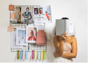 Picture of a girl hiding his face with a book standing next to several fashion magazines